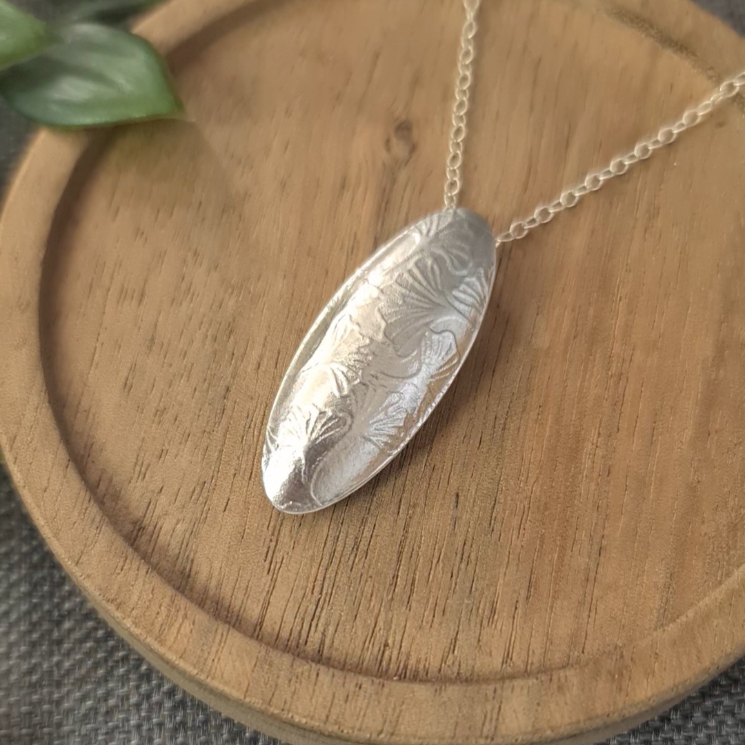 image shows the finished project, a hollow pendant with a ginkgo leaf pattern embossed into the silver. The pendant is hanging from a chain and is laying on a small round wooden plate.